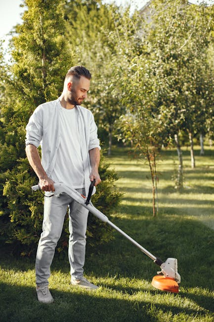 A man with a neatly groomed beard and styled hair is wearing a light grey shirt with rolled-up sleeves and light grey trousers while operating a modern, electric grass trimmer to edge a well-maintaine